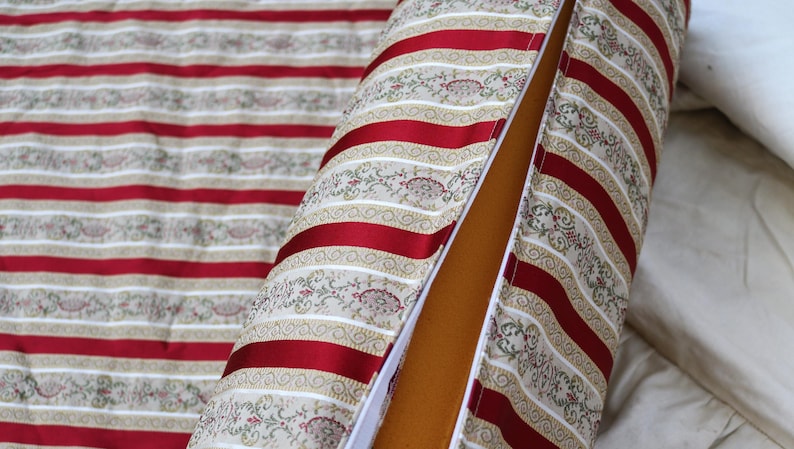 May include: A cylindrical pillow and matching fabric with a red and beige striped pattern. The pillow has a red stripe design with floral accents and a zipper. The interior is a solid tan color.