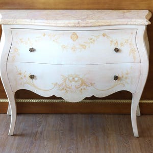 Può includere: Una cassettiera in legno color crema con piano in marmo, dotata di due cassetti decorati con delicati motivi floreali dipinti in oro e quattro piccoli pomelli in metallo scuro. Il mobile presenta fianchi curvi e poggia su quattro gambe sottili.