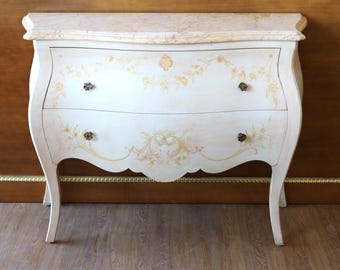 May include: A cream-colored wooden chest of drawers with a marble top, featuring two drawers adorned with delicate gold floral painted motifs and four small dark metal knobs. The piece has curved sides and stands on four slender, flared legs.