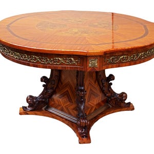 Può includere: Un tavolo rotondo in legno con un piano in marquetry e una base scolpita. Il tavolo ha un bordo in ottone attorno al bordo del piano e un design decorativo sulla base.