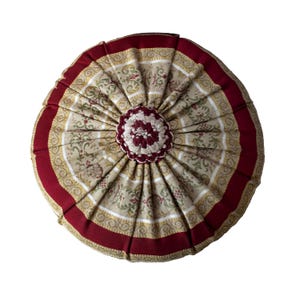 May include: Round decorative pillow with a floral pattern in beige, red, and gold. The pillow has a pleated design with a dark red border and a central, flower-shaped embellishment. The pillow is approximately 16 inches in diameter.