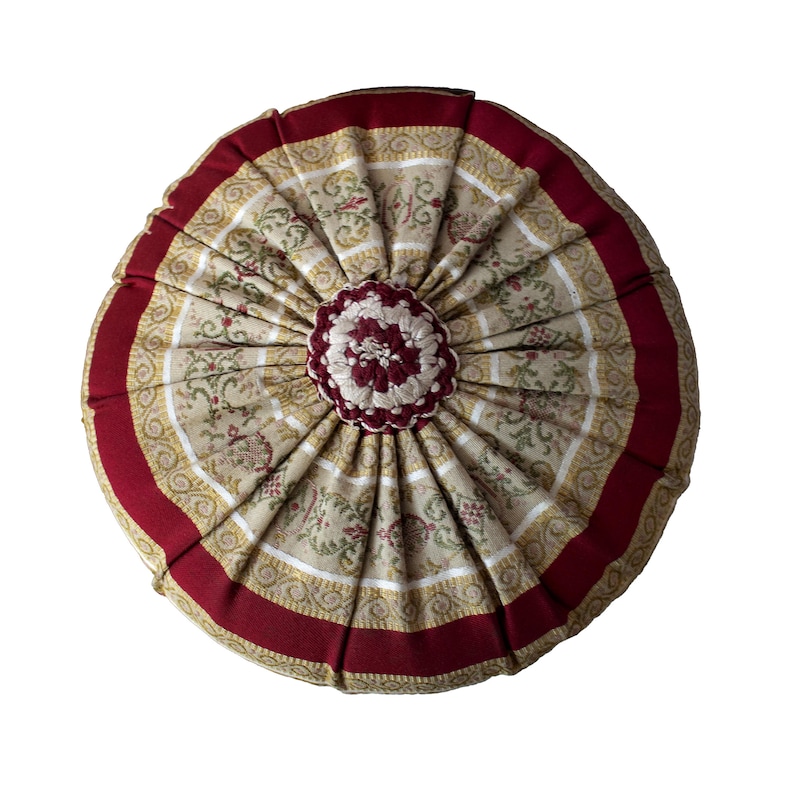 May include: Round decorative pillow with a floral pattern in beige, red, and gold. The pillow has a pleated design with a dark red border and a central, flower-shaped embellishment. The pillow is approximately 16 inches in diameter.