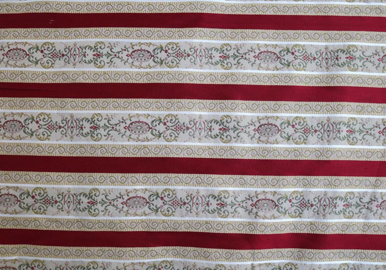 May include: Close-up of a fabric with a repeating pattern of alternating red stripes and decorative beige bands. The beige bands feature floral and scrollwork designs in green, red, and gold.