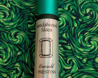 May include: A cylindrical perfume bottle with a teal cap and a silver label. The label features the words "sandalwood Skies" above a diamond-shaped graphic and the words "Emerald Priestess" below. The background is a green, swirling pattern.