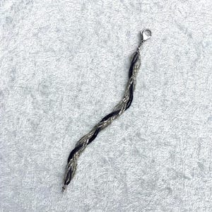 May include: Braided bracelet featuring black and silver strands. The bracelet is made of small, shiny beads woven together in a textured pattern. It has a silver-tone clasp for closure. The bracelet is displayed on a white, textured surface.