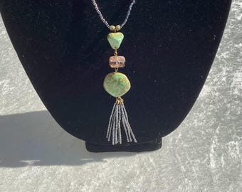 Op de afbeelding: Een ketting met een kralenketting en een hanger met een hartvormige turkooizen steen, een roze rechthoekige kraal en een grotere turkooizen steen. De hanger eindigt met een kwastje van kleine grijze kralen. De ketting wordt getoond op een zwarte mannequin.
