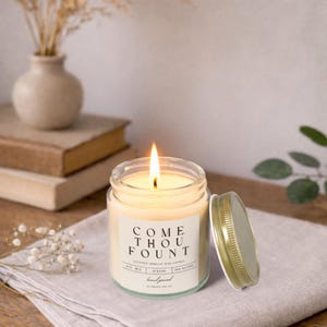 Puede incluir: Una vela encendida en un tarro de cristal transparente con tapa dorada. La vela tiene cera color crema y una etiqueta que dice "COME THOU FOUNT". La vela está sobre una tela blanca, con libros y un jarrón en el fondo.
