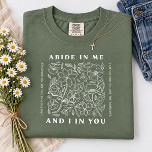 May include: An olive green t-shirt with a white graphic print featuring a floral design and the text "ABIDE IN ME AND I IN YOU" along with "I AM THE VINE YOU ARE THE BRANCHES" written vertically on the sides of the central flower illustration.