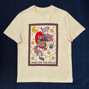 Puede incluir: Camiseta color crema con un gráfico colorido. El gráfico presenta un caballero montando a caballo, sosteniendo una bandera con un diseño de bandera del orgullo transgénero. El texto "I RIDE FOR THE DOLLS" está en la parte inferior del gráfico.
