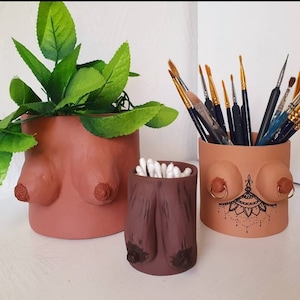 May include: Three terracotta-coloured ceramic containers shaped like breasts. One holds a plant, another holds cotton buds, and the third holds paintbrushes. The third container has a decorative tattoo design and gold-coloured rings.