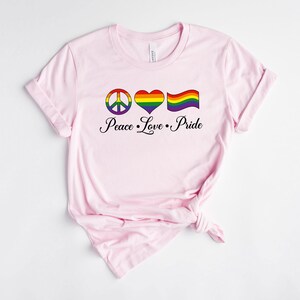 Op de afbeelding: Roze T-shirt met een vredesteken, hart en regenboogvlag. De tekst "Peace Love Pride" staat onder de afbeelding.