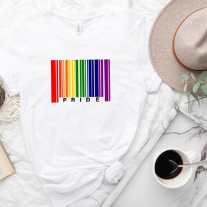 Peut inclure: T-shirt blanc avec un motif de code-barres coloré aux couleurs de l'arc-en-ciel. Le mot "PRIDE" est imprimé en dessous. Le t-shirt est présenté sur une surface blanche avec un livre, du café et un chapeau.