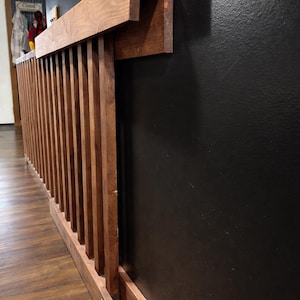 K&ouml;nnte beinhalten: Ein h&ouml;lzernes Gel&auml;nder mit vertikalen Lamellen und einem horizontalen Handlauf, befestigt an einer schwarzen Wand. Das Gel&auml;nder besteht aus gebeiztem Holz, und das Design ist modern und minimalistisch. Das Holz hat einen warmen, nat&uuml;rlichen Farbton.