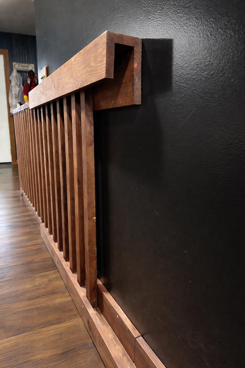 K&ouml;nnte beinhalten: Ein h&ouml;lzernes Gel&auml;nder mit vertikalen Lamellen und einem horizontalen Handlauf, befestigt an einer schwarzen Wand. Das Gel&auml;nder besteht aus gebeiztem Holz, und das Design ist modern und minimalistisch. Das Holz hat einen warmen, nat&uuml;rlichen Farbton.