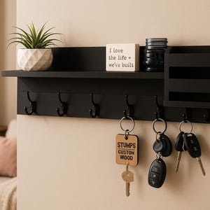 Puede incluir: Soporte de llaves de madera negra con un estante, ganchos y un organizador de correo. Una pequeña planta en maceta, un bloque decorativo con texto y llaves cuelgan de los ganchos. El texto en el bloque dice "I love the life we've built."