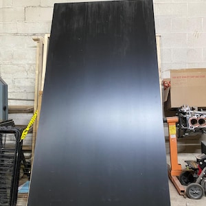 May include: A large, rectangular black panel leans against a brick wall. The panel has a smooth, matte finish with subtle vertical streaks. The panel is likely a component for construction or a similar project.