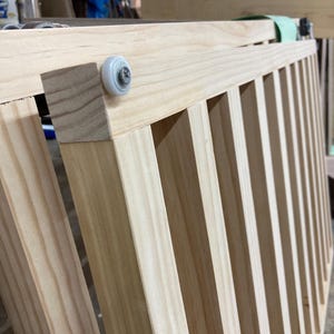 K&ouml;nnte beinhalten: Nahaufnahme einer Holz-Kinderbettseite mit vertikalen Sprossen. Das unbehandelte Holz zeigt nat&uuml;rliche Maserungsmuster. Eine wei&szlig;e Rolle ist an der Ecke befestigt, was auf einen Schiebemechanismus hindeutet. Die Kinderbettseite befindet sich in einer Werkstatt.