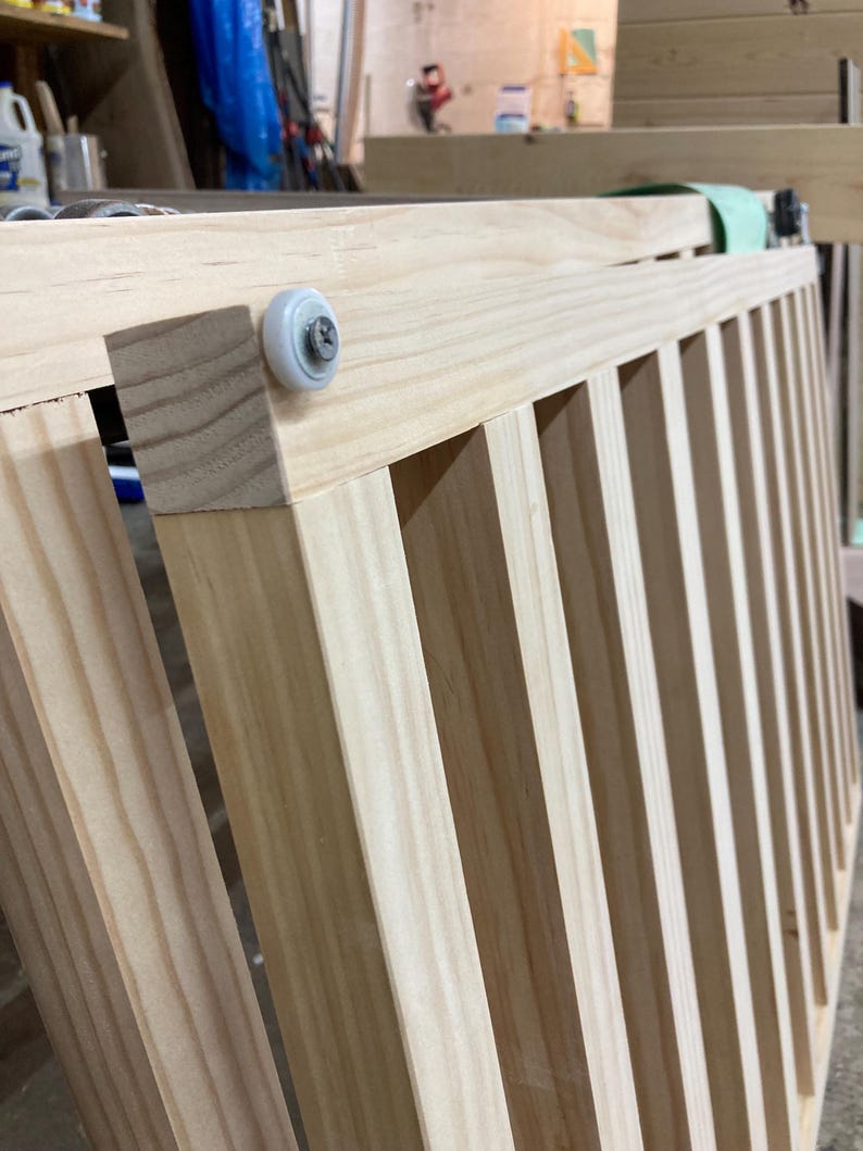 K&ouml;nnte beinhalten: Nahaufnahme einer Holz-Kinderbettseite mit vertikalen Sprossen. Das unbehandelte Holz zeigt nat&uuml;rliche Maserungsmuster. Eine wei&szlig;e Rolle ist an der Ecke befestigt, was auf einen Schiebemechanismus hindeutet. Die Kinderbettseite befindet sich in einer Werkstatt.