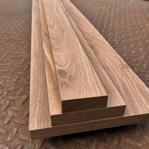Op de afbeelding: Drie rechthoekige houten planken die op elkaar zijn gestapeld. De planken hebben een natuurlijk bruin houtnerfpatroon met zichtbare lineaire texturen en gladde, vlakke oppervlakken. Elke plank heeft rechte, strak gesneden randen.