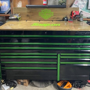 May include: Black and green Snap-on tool chest with multiple drawers. The top of the chest has a wooden surface with the words "Snap-on" and "Motor Harley Davidson Cycles" printed on it.
