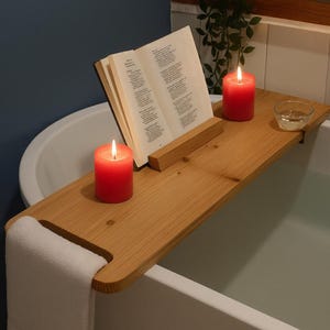 Puede incluir: Una bandeja de bañera de madera con un libro, dos velas rojas y un pequeño cuenco de cristal. Una toalla blanca cuelga de la bandeja. Las velas están encendidas y el libro está abierto. La bandeja está sobre una bañera blanca.