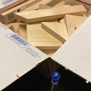 May include: A cardboard box filled with various sizes and shapes of unfinished wooden pieces. The box is open and a blue marker is visible in the bottom of the box.