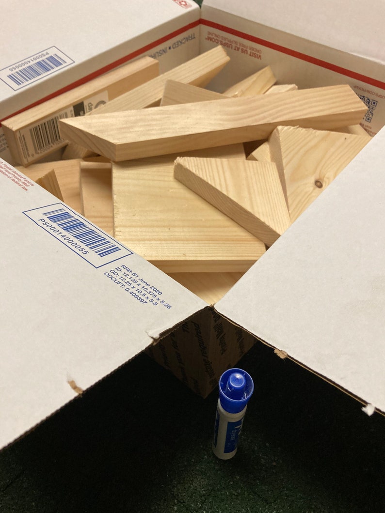 May include: A cardboard box filled with various sizes and shapes of unfinished wooden pieces. The box is open and a blue marker is visible in the bottom of the box.