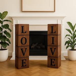 May include: Two sets of wooden blocks spelling "LOVE" and "LIVE". The blocks are dark brown with black letters. The blocks are stacked in a vertical arrangement. The background includes a fireplace, plants, and a framed picture.