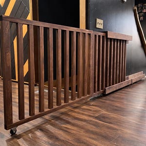 K&ouml;nnte beinhalten: Dunkelbraunes Babygitter aus Holz mit vertikalen Lamellen und einem Rollrad unten. Das Gitter ist in einem Raum mit dunklem Holzboden und einer schwarzen Wand installiert.