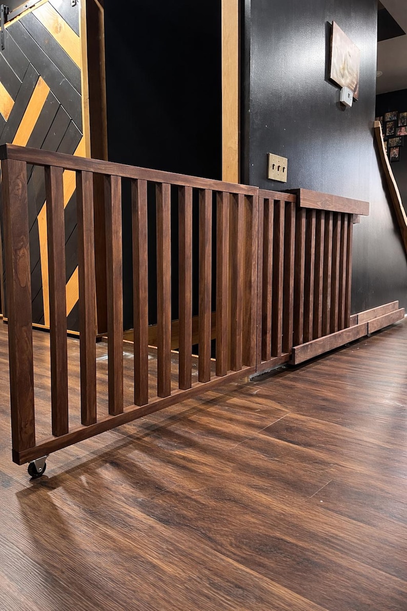 K&ouml;nnte beinhalten: Dunkelbraunes Babygitter aus Holz mit vertikalen Lamellen und einem Rollrad unten. Das Gitter ist in einem Raum mit dunklem Holzboden und einer schwarzen Wand installiert.