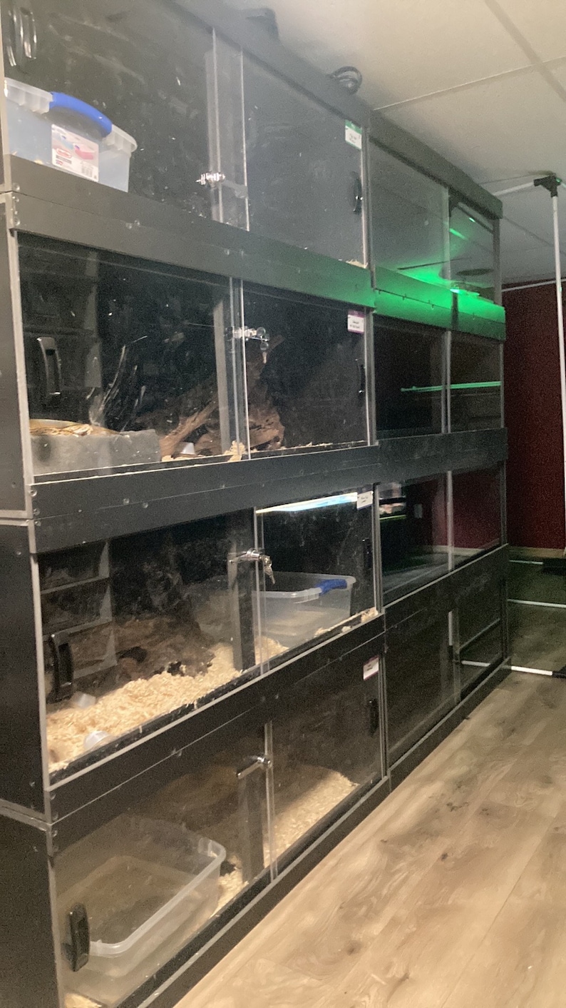 May include: A stack of black reptile cages with glass doors and clear plastic containers inside. The cages are stacked on top of each other and have a wooden floor.