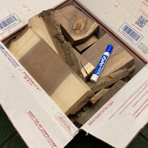 May include: A cardboard box filled with various pieces of wood, including a few with knots. A blue dry-erase marker is lying on top of the wood pieces. The box is labelled "PRIORITY MAIL" and "VISIT US AT USPS.COM".
