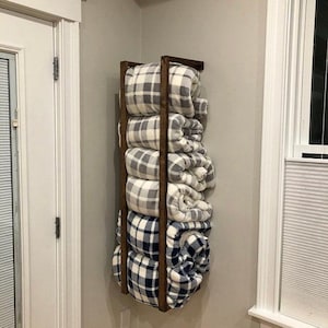 May include: A wooden wall-mounted blanket storage rack with five rolled blankets in a black and white plaid pattern. The rack is made of dark brown wood and has a rustic look.