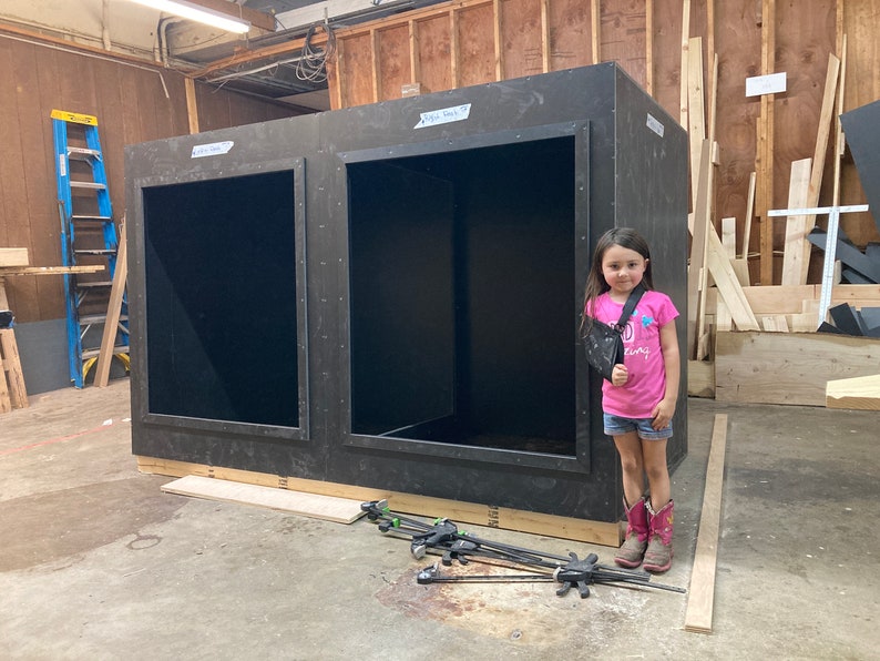 May include: A young girl wearing a pink shirt and denim shorts stands in front of a large wooden structure with two open doorways. The structure is made of plywood and has a black interior. The girl is wearing a sling on her left arm.