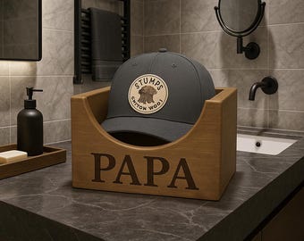 May include: A gray baseball cap with a circular patch that reads "STUMPS CHITON WOOL" sits in a wooden box. The box is engraved with the word "PAPA". The hat and box are on a dark countertop in a bathroom setting.