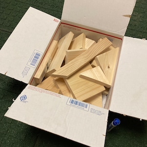 May include: A cardboard box filled with various sizes and shapes of unfinished wooden pieces. The box is open and the wooden pieces are visible inside. The box has a label with a barcode and the text "PS00914000055".