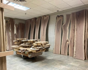 Op de afbeelding: Een werkplaats met een grote collectie houten platen, sommige gestapeld op een kar. De platen zijn van verschillende groottes en vormen, met een natuurlijk houtnerfpatroon. Het hout is lichtbruin.