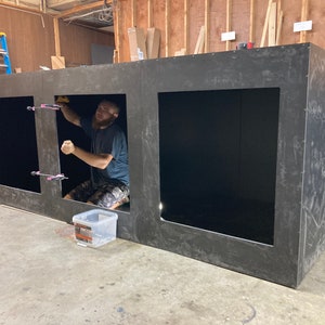 May include: A person is working on a large, black, rectangular structure with three open square compartments. The structure is made of a thick, dark material and appears to be unfinished. The person is wearing a blue shirt and is using tools to secure the structure.