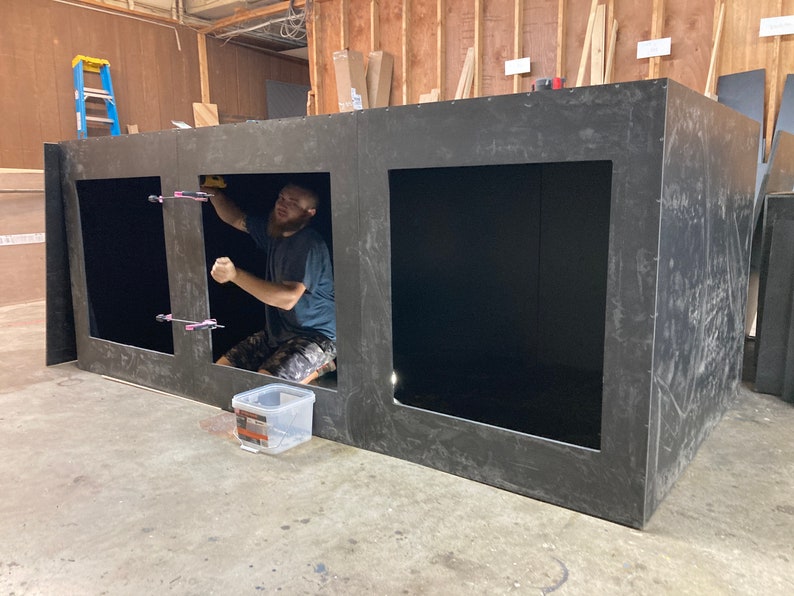 May include: A person is working on a large, black, rectangular structure with three open square compartments. The structure is made of a thick, dark material and appears to be unfinished. The person is wearing a blue shirt and is using tools to secure the structure.