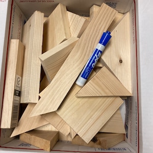 May include: A cardboard box filled with various sizes and shapes of unfinished wood pieces. The box is labeled "Regional Rate Box B" and "Tracked - Insured". A blue dry-erase marker is visible on top of the wood pieces.
