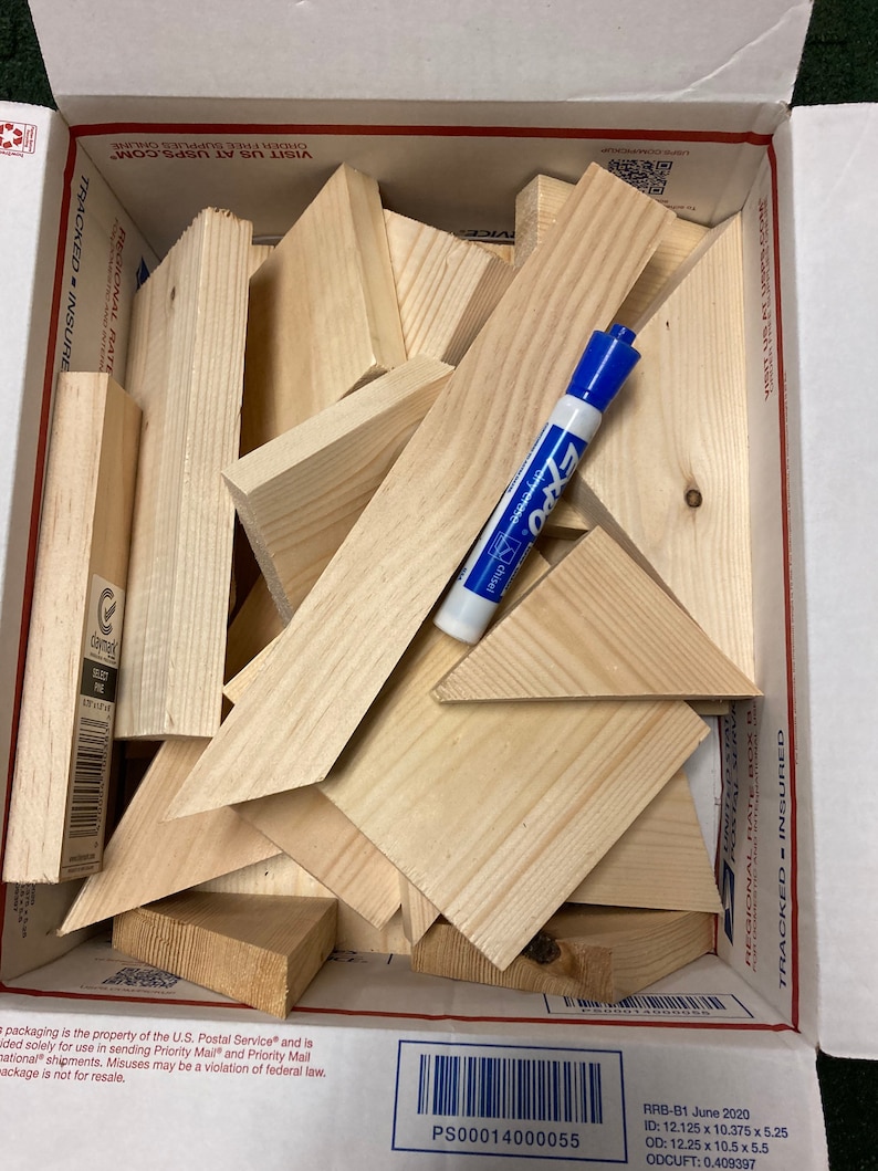 May include: A cardboard box filled with various sizes and shapes of unfinished wood pieces. The box is labeled "Regional Rate Box B" and "Tracked - Insured". A blue dry-erase marker is visible on top of the wood pieces.