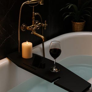 May include: A black bath tray with a lit candle, a smartphone, and a glass of red wine. The tray sits on the edge of a white bathtub filled with water. A gold tap and showerhead are in the background.