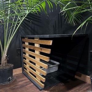 May include: A black wooden cat litter box enclosure with a slatted wooden front. The enclosure has a rectangular shape and a dark interior. A gray litter box is visible inside. The background features green plants and a black wall.