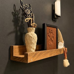 May include: A rectangular wooden floating shelf holding a cream-colored textured vase with wooden flower cutouts, a framed brown square featuring an embossed paw print, and a small gnome figurine with a tan hat and dangling fabric legs.