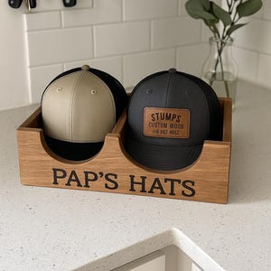 Könnte beinhalten: Ein hölzerner Hutständer mit dem schwarzen Schriftzug "PAP'S HATS". Er enthält zwei Baseballkappen: eine beige und schwarze, die andere dunkelgrau mit einem Lederaufnäher "STUMPS CUSTOM WOOD".