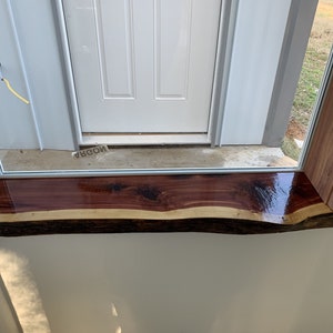 May include: A close-up of a windowsill made of a natural wood slab. The wood is a rich reddish-brown color with a natural edge and a clear coat finish. The windowsill is installed above a white door.