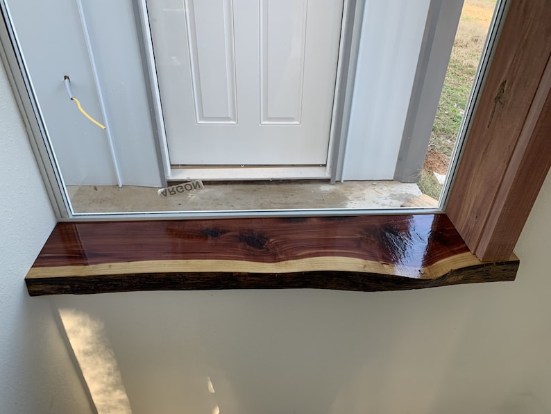 May include: A close-up of a windowsill made of a natural wood slab. The wood is a rich reddish-brown color with a natural edge and a clear coat finish. The windowsill is installed above a white door.
