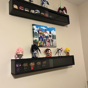 Könnte beinhalten: Zwei schwarze, rechteckige Wandvitrinen mit Glasfronten, die jeweils mehrere kleine Vinyl-Figuren enthalten. Über und unter den Vitrinen befinden sich weitere Figuren, und ein rechteckiger Leinwanddruck zeigt eine Gruppe von fünf Personen.