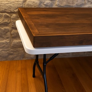 Könnte beinhalten: Eine rechteckige Tischplatte aus Holz mit dunkelbraun gebeizter Oberfläche und sichtbarer Holzmaserung, die auf einem weißen Klapptisch aus Kunststoff liegt, welcher schwarze Metallbeine mit geschwungenen Stützen und Gummifüßen besitzt.
