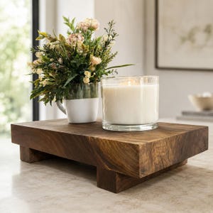 May include: A rectangular wooden riser with four short feet, supporting a white ceramic mug filled with a bouquet of pink and green flowers and a clear glass jar containing a white candle with a visible flame, creating a decorative display.
